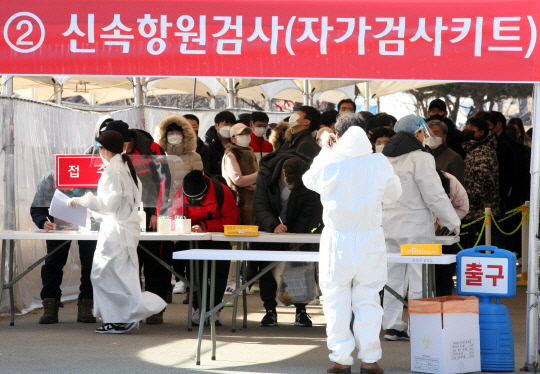 韩国检疫站，医护人员身穿防护服正在协助市民进行自我检测，背景为正在排队等待检测的人群。这张图片描述了COVID-19疫情期间的检测情况。